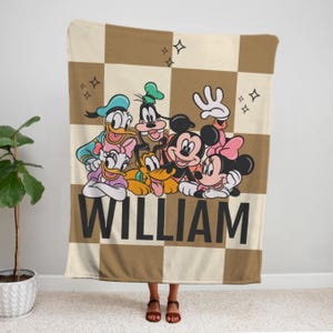 Puede incluir: Una manta a cuadros marr&oacute;n y beige con personajes de dibujos animados de Disney, como Mickey y Minnie Mouse, Donald y Daisy Duck, Goofy y Pluto. El nombre "WILLIAM" est&aacute; impreso en letras negras grandes.