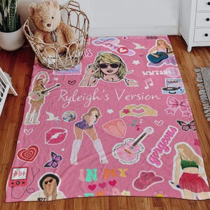 May include: Pink blanket with illustrations of a woman, musical instruments, hearts, and the text "Ryleigh's Version". The blanket has a variety of graphics and the word "gorgeous".