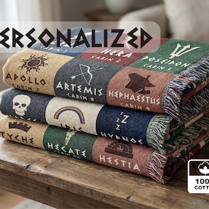 May include: A stack of personalized woven blankets with various designs and text, including "PERSONALIZED." Each blanket features different colors and graphics, such as a sun, skull, and rainbow. The blankets are made of 100% cotton and have fringed edges.