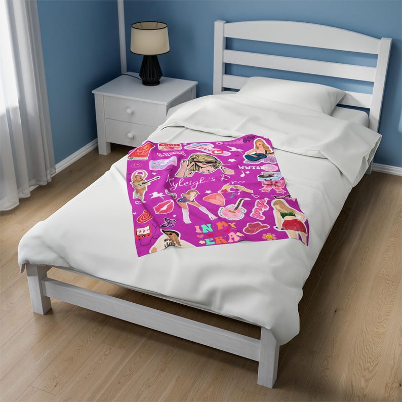 May include: A magenta blanket with a collage of illustrations and text, including the words "Tyleigh's Era" and "In My Era." The blanket features images of a woman, musical instruments, and other graphics. The blanket is on a white bed.