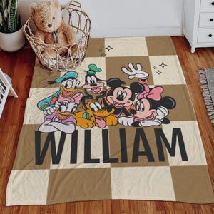 Puede incluir: Una manta a cuadros marr&oacute;n y beige con personajes de dibujos animados de Disney, incluyendo Mickey y Minnie Mouse, Donald y Daisy Duck, Goofy y Pluto. El nombre "WILLIAM" est&aacute; impreso en letras negras grandes.