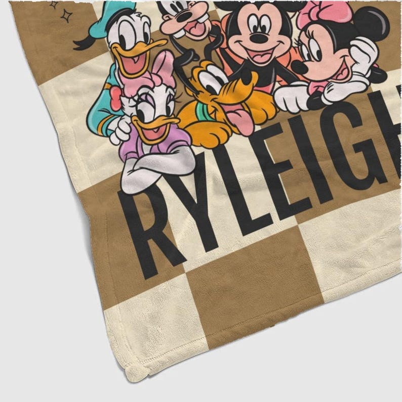 Puede incluir: Una manta a cuadros beige y crema con personajes de Disney, incluyendo Mickey y Minnie Mouse, Donald y Daisy Duck, y Pluto. El nombre "RYLEIGH" est&aacute; impreso en letras negras grandes.