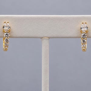 May include: A pair of gold hoop earrings featuring a row of clear, round-cut stones and a single pear-cut stone at the top. The earrings are displayed on a horizontal white stand, showcasing the metallic finish and the sparkling stones.