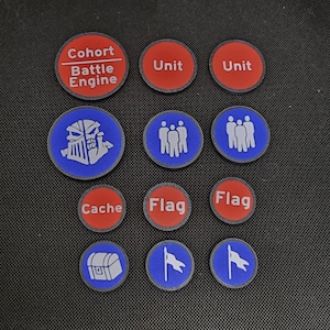 May include: A collection of circular game tokens with various icons and text. The tokens are primarily red and blue with white or gray graphics. Text includes "Cohort Battle Engine", "Unit", "Cache", and "Flag".