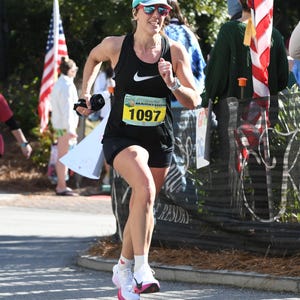 Puede incluir: Una mujer con un top negro sin mangas, pantalones cortos negros, un sombrero blanco y gafas de sol corre un maratón. Su número de carrera es 1097. La carrera se llama "Coastal Marathon".