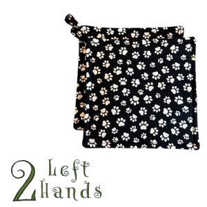Puede incluir: Dos agarraderas con estampado de huellas de patas en blanco y negro, con un lazo para colgar. Las agarraderas cuadradas presentan un patrón repetido de huellas de patas blancas sobre un fondo negro. El texto "2 Left Hands" está en la parte inferior de la imagen.