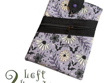 Könnte beinhalten: Eine rechteckige, lavendelfarbene Tasche mit einem schwarzen Reißverschluss und Klappenverschluss. Die Tasche hat ein Blumen- und Fledermausmuster. Der Text "2 Left Hands" ist unten in grüner Schrift zu sehen.