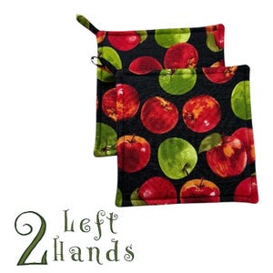 Puede incluir: Dos agarraderas negras con un estampado de manzanas rojas y verdes. Las agarraderas tienen un lazo para colgar. El texto "2 Left Hands" está en verde en la parte inferior de la imagen.
