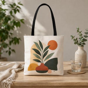 May include: Cream-colored tote bag with a black handle featuring an abstract design. The design includes a terracotta vase with green leaves, an orange sun, and geometric shapes in yellow and dark green. The bag is on a wooden table.