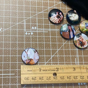 Peut inclure: Un ensemble de six ornements photo ronds en verre avec diff&eacute;rentes images. Les ornements mesurent environ 2,5 cm de diam&egrave;tre et sont pr&eacute;sent&eacute;s sur un tapis de coupe brun avec une r&egrave;gle.
