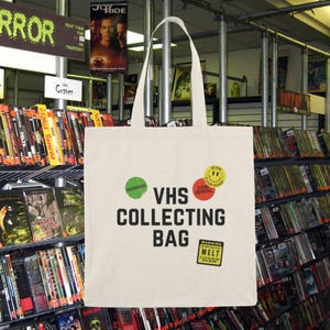 May include: A white canvas tote bag with the text "VHS COLLECTING BAG" in black. The bag has three stickers: a green sticker with the word "HORROR", a red sticker with the text "3 DAY RENTAL", and a yellow sticker with a smiley face. The bag is in front of a shelf full of VHS tapes.