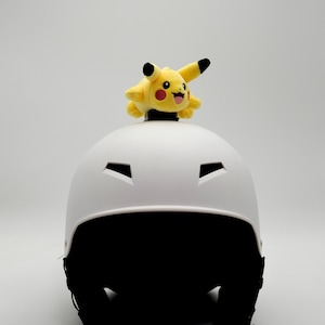 May include: A white helmet with black accents and a yellow Pikachu plush toy on top. The plush toy has black ears and a red mouth. The helmet has several ventilation openings.