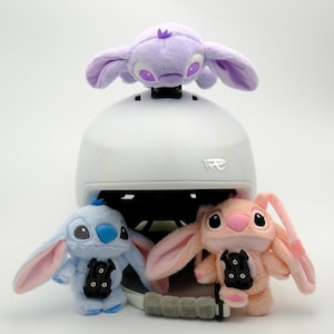 May include: A white helmet with three plush toys of a cartoon character. The toys are in light blue, pink, and purple. The toys have large ears and black accents. The helmet has a logo on the front.