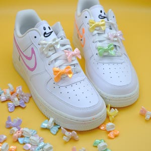 May include: White sneakers with colorful accents, including a pink Nike swoosh and smiley face details. The laces are adorned with pastel-colored bow charms. A playful, fashionable footwear design.