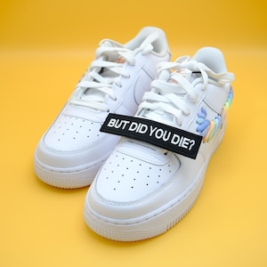 May include: Pair of white sneakers with a black tag that reads "BUT DID YOU DIE?" The shoes have a classic design with white laces and a colorful accent on the side. The background is a solid yellow.