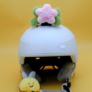 May include: A white ski helmet with a plush pink and green flower accent on top. A small, smiling plush bee is attached to the helmet's chin strap. The helmet is on a yellow background.