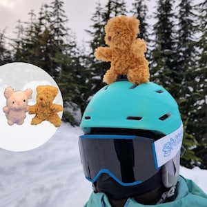 May include: A person wearing a blue and white ski helmet with a brown teddy bear on top. The helmet has a blue tinted visor and a white strap with the word "Smith" on it. The person is wearing a blue and green jacket. There are two smaller teddy bears in the background.