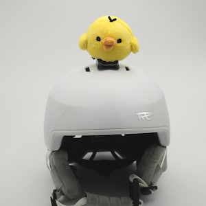 May include: A white ski helmet with a plush yellow chick perched on top. The helmet has gray padding and a chin strap. The chick has a black eye and a small orange beak. The helmet is designed for winter sports.