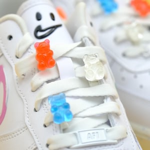 May include: White leather trainers with gummy bear charms in blue, orange, and white. The trainers have a smiley face on the side and the text "AF1" on the tongue.