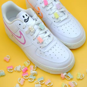 May include: White trainers with colourful gummy bear shoe charms on the laces. The trainers have a pink and orange swoosh logo. There are several gummy bear charms scattered around the trainers.