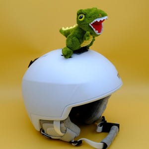 May include: A white ski helmet with a plush green dinosaur attached to the top. The dinosaur has a red mouth and yellow eyes. The helmet has a chin strap and ear padding. The background is a solid yellow.