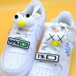 May include: White sneakers with custom shoe charms. One shoe has a fluffy white charm with black eyes and a yellow nose, and a patch that reads "99%" with a battery icon. The other shoe has a similar charm and a patch that reads "1%" with a battery icon.