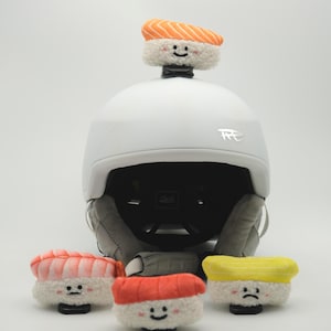 May include: A white ski helmet with a sushi-themed design. The helmet has a sushi topping on top and several sushi-shaped accessories at the base. The sushi toppings include salmon, shrimp, and egg, each with a smiling or sad face.