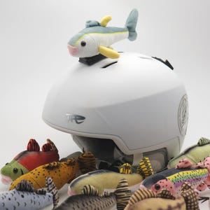 May include: A white helmet with a plush fish attached to the top, surrounded by several colorful plush fish toys. The fish toys have various colors and patterns, including yellow, green, and red. The helmet has a logo on the side.