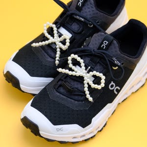 May include: A pair of black and white sneakers with pearl-beaded bows tied on the laces. The shoes have a white sole and a black upper with a mesh design.
