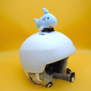 May include: A white ski helmet with a blue plush shark attached to the top. The shark has a black eye and a red mouth.