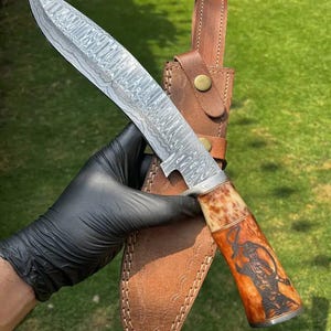 May include: A Damascus steel hunting knife with a brown and tan handle featuring an etched design. The knife is displayed with a brown leather sheath. The blade has a unique pattern. The handle has a decorative design.