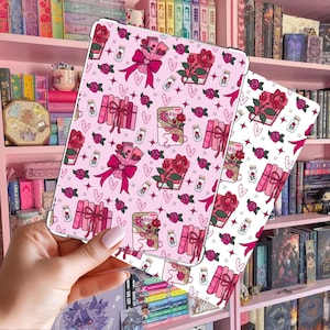 Könnte beinhalten: Zwei rosa Tablet-Hüllen mit einem sich wiederholenden Muster aus Rosen, Büchern, Blumensträußen und Gläsern. Die Hüllen werden vor einem rosa Bücherregal mit Büchern gehalten. Das Muster enthält Herzen und Sterne.