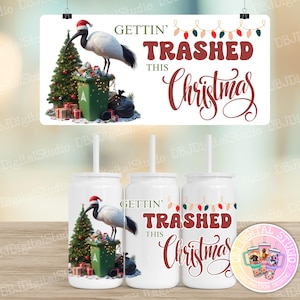May include: White tumbler with a straw featuring a Christmas design. The design includes a bird wearing a Santa hat standing on a trash can next to a Christmas tree and the text "GETTIN' TRASHED THIS Christmas".