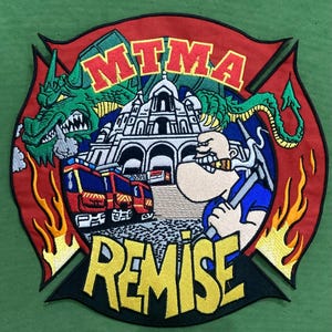 Puede incluir: Un parche bordado circular con un borde de cruz de Malta roja y llamas amarillas. El diseño incluye un dragón verde, un edificio blanco, camiones de bomberos rojos y un personaje de dibujos animados con el texto MTMA y REMISE.