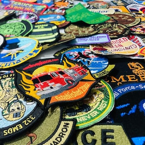 May include: A large collection of assorted embroidered fabric patches featuring various shapes, colors, and designs. Visible text includes '332 EMDS', 'MEDICAL SQUADRON', 'TOWN OF TROY', 'BEARDED MAN UNSHAVED', 'ESTABLISHED 2002', and 'FIREAPPLIANCES.COM'.