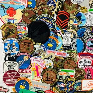 Op de afbeelding: Een dichte verzameling geborduurde patches met diverse vormen, kleuren en ontwerpen. Zichtbare tekst bevat Mr. Rooter, Huntington Beach, Yacht Water, Mobstar, KSMO, Railhouse Pub, Valkyrie, Kratos en Evolution Power Solutions.
