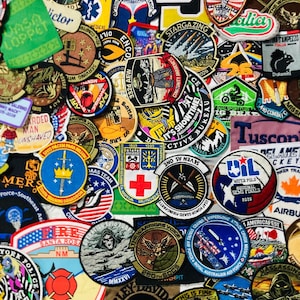 May include: A collection of numerous embroidered fabric patches featuring various shapes, colors, and designs, including military emblems, sports logos, text, and graphic illustrations, layered together in a dense, overlapping arrangement.
