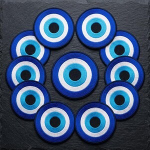 Può includere: Una collezione di dodici toppe ricamate circolari con un design concentrico a occhio di Allah. Ogni toppa presenta una pupilla nera centrale circondata da anelli di colore azzurro, bianco e un bordo esterno di colore blu scuro.