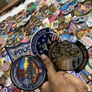 Pode incluir: Uma coleção de emblemas de tecido bordado com vários designs, incluindo um emblema circular com uma aeronave militar e o texto OP REASSURANCE ATF-P, um emblema GVIII ROAAS TEAM e um emblema 332 EMDS PHARMACY com uma figura de desenho animado.