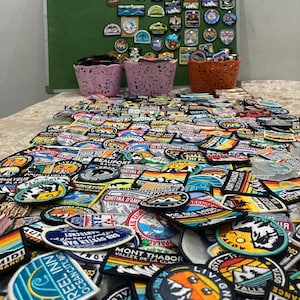 Op de afbeelding: Een grote collectie geborduurde souvenirpatches met diverse berglandschappen, reisbestemmingen en logo's. Veel patches zijn bevestigd op een groen prikbord, terwijl andere op een tafel voor drie kleine mandjes liggen opgestapeld.