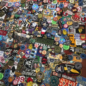 May include: A collection of embroidered patches in various shapes and sizes, featuring text, logos, and designs. The patches display a range of colors, including red, blue, green, and yellow. Some patches have military, sports, and commercial themes.