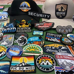 Op de afbeelding: Een verzameling van diverse geborduurde patches met uiteenlopende ontwerpen zoals berglandschappen, dieren en tekst, samen met een zwarte baseballpet met een eendafbeelding en een beige baseballpet met een bergpatch.
