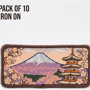Puede incluir: Un parche termoadhesivo rectangular con un paisaje bordado del monte Fuji y una pagoda de varios niveles rodeada de cerezos en flor en tonos rosa, morado y marrón. El texto PACK OF 10 IRON ON aparece impreso en la esquina superior izquierda.