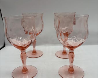 May include: Four pink-tinted wine glasses with textured bowls and delicate stems. The glasses have a tulip-like shape and sit on round bases. The glassware is made of a transparent material.