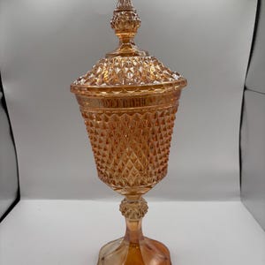 May include: An iridescent orange glass candy dish with a lid. The dish has a diamond pattern and sits on a pedestal base. The lid is topped with a decorative finial. The glass has a shimmering, reflective quality.