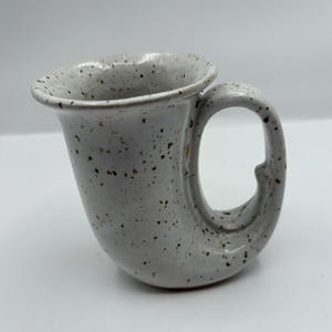 Puede incluir: Una taza de cerámica moteada de color blanquecino con un asa curva única. La taza tiene un borde acampanado y un patrón moteado de puntos marrones y negros. El asa se curva con elegancia, lo que se suma al diseño distintivo de la taza. Es una pieza hecha a mano.