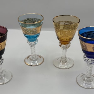 Puede incluir: Juego de cuatro vasos de chupito vintage de vidrio con ribete dorado y vidrio de diferentes colores. Los vasos son transparentes, azules, morados y ámbar.