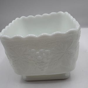 May include: A white milk glass square bowl with a scalloped rim. The sides feature a detailed embossed grape and leaf design. The bowl sits on a small, rectangular base. This vintage piece is ideal for serving or display.