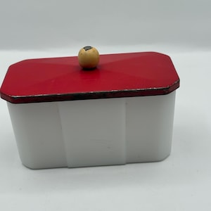 May include: A rectangular white container with a red lid. The lid has a small, round, wooden knob on top. The container has a geometric design and is set against a white background.
