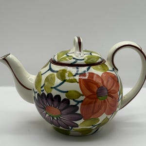 Puede incluir: Tetera de cerámica color crema con diseño floral. La tetera tiene un cuerpo redondo, un asa curva y un pico. El patrón floral incluye flores y hojas naranjas, moradas y verdes. La tapa tiene un asa pequeña.